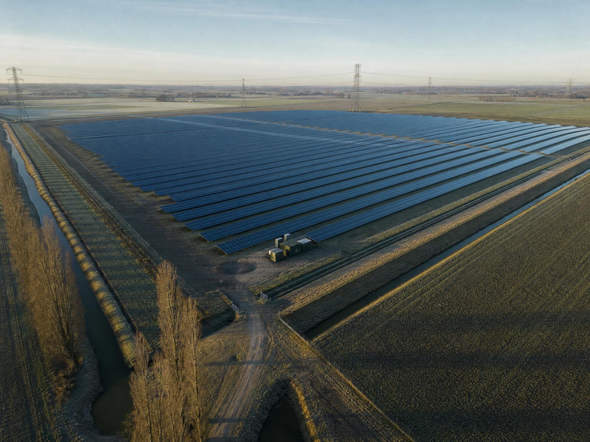 Luchtfoto van een zonnepark in een vlak landschap, met een hoogspanningslijn aan de horizon