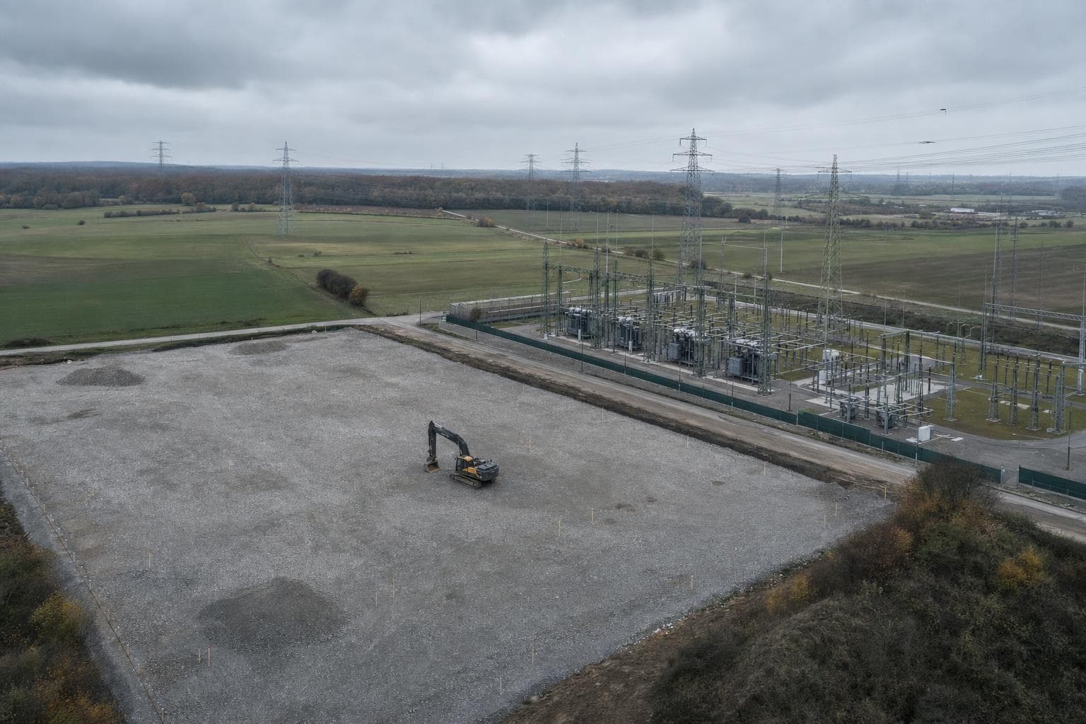 Luchtfoto van een verhard, leeg terrein met piketpaaltjes en een graafmachine, direct ernaast een onderstation met hoogspanningslijnen